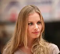 Jana Beller mit langen, blonden Haaren und grünen Augen, lächelt leicht in einem hellen Oberteil. Im Hintergrund könnten Gebäude oder Strukturen zu erkennen sein, aber die Details sind nicht klar. Jana Beller scheint aufmerksam einem Reporter oder Gesprächspartnern zuzuhören.