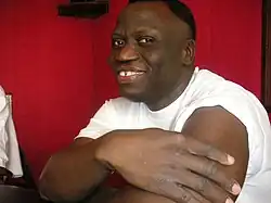 Jean Odoutan. 2005. Ein sitzender afrikanischer Mann, der die Hände verschränkt und auf den Tisch stützt.