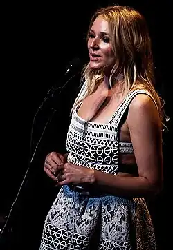 Jewel steht vor einem schwarzen Mikrofon, in das sie reinsingt. Sie hat blonde, schulterlange Haare und trägt ein weißes Schürzenkleid, auf dem mehrere schwarze Muster, unter anderem Rauten, Rechtecke und Schnörkel, zu sehen sind.