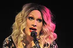 Monsoon steht vor einem Mikrofon, in das sie reinspricht. Sie trägt eine blonde Perücke, falsche Wimpern, aufgemalte Augenbrauen, roten Lippenstift, eine Halskette und ein schwarzes Kleid mit Blumenmuster.