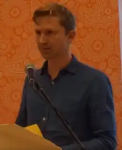 Ein verpixeltes Bild, zu sehen ein blonder Mann im Hemd bei einer Lesung.