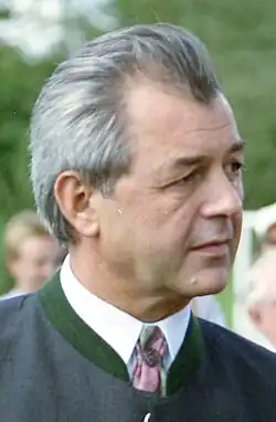 Brustporträt eines Mannes mit grauem, nach hinten gekämmtem Haar, einen Trachtenjanker tragend, rechts aus dem Bild hinausschauend