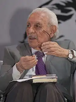 Ein älterer Mann, sitzend, spricht in der Öffentlichkeit.