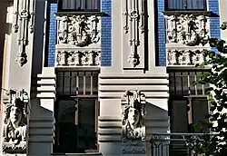 Farbige Nahaufnahme eines Fassadenteils mit Kopfreliefs an beiden Fensterseiten und Ornamenten über den Fenstern.