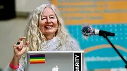 Jutta Hartmann ist mit vor einem unscharfen Hintergrund zu sehen, während sie vor einem Laptop sitzt und den rechten Arm halb erhoben zu einer Geste positioniert hat. Sie ist während des Sprechens zu sehen und hat lange helle gelockte Haare.