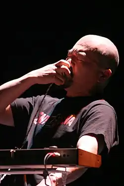 Der kahl rasierte Musiker hält sich, mit geschlossenen Augen, ein Mikrofon vor den Mund. Eine Kante der Rückseite des Synthesizers ist im Vordergrund zu sehen.
