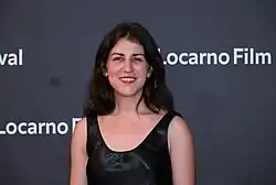 Katharina Lüdin mit langen, dunklen Haaren und rotem Lippenstift, trägt ein ärmelloses schwarzes Kleid, lächelt auf dem roten Teppich des 76. Locarno Film Festival 2023. Sie steht vor einem Hintergrund, auf dem wiederholt der Text „Locarno Film Festival“ zu sehen ist.
