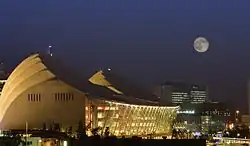 Das Kauffman Center for the Performing Arts bei Nacht. Zu sehen ist die gut beleuchtete, leicht abgeschrägte Glasfront in der Bildmitte. Die abgerundeten Gebäudeflügel, die links im Bild zu sehen sind, sind auf ihrer Rückseite ebenfalls gut beleuchtet. Im Hintergrund sieht man den Vollmond.