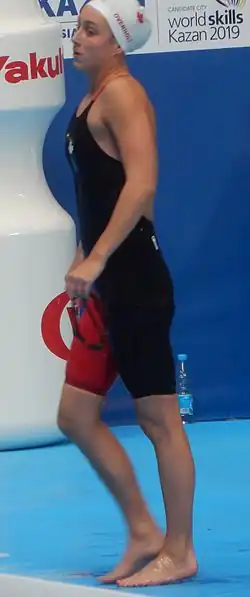 Emily Overholt trägt einen dunklen Badeanzug, bei dem ein Bein rot ist, und eine weiße Badekappe, auf der ein rotes Ahornblatt erkennbar ist und ihr Nachname in roter Schrift. In der linken Hand hält sie ihre Schwimmbrille. Im Hintergrund befindet sich eine blaue Wand mit Werbeaufschriften in lateinischer Schrift.