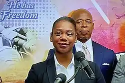 Zwei Personen vor einem mehrfarbigen Hintergrund mit dem aufgedruckten Claim „He has Freedom“. Vorne an einem Mikrofon lächelnd Keechant Sewell, direkt rechts hinter ihr Eric Adams.