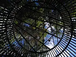 Innerhalb einer runden Stahlkonstruktion, von unten in der Mitte fotografiert, hängen oben verteilt über einem Metallgitter die Porzellanglocken unter freiem Himmel.