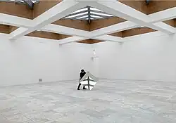 Ein Mann richtet in einem leeren Ausstellungsraum einen mehr als einen Quadratmeter großen Spiegel so aus, dass dieser einen Teil der Dachkonstruktion zeigt.