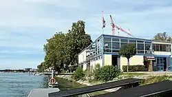 Zusehen ist das Ruderhaus mit den Bootshallen unten und dem Restaurant oben. Im Vordergrund sind die Stege zu sehen, über die die Boote zum See gelangen.