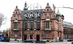 Frontale Farbfotografie eines zweigeschossigen, braunen Gebäudes mit zwei Staffelgiebeln an den Ecken. Über dem Eingang befindet sich ein Giebelfeld, neben dem an beiden Seiten der Name „Tivoli“ steht. Dieser Name steht in großen Buchstaben auf einem Dachgerüst. Im Obergeschoss sind drei Rundbogenfenster von zwei Fenstervorbauten gesäumt. An der Seite sind eine Turmhaube und ein Reklamegerüst zu sehen. Im Hintergrund ist eine Turmspitze zu erkennen.