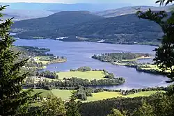 Foto von einer Anhöhe auf einen See herab, der in einer hügeligen und teils bewaldeten Umgebung liegt