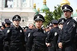 Im Vordergrund der Farbfotografie stehen drei Polizisten, jeweils ein Mann links und rechts vom Foto und eine Frau in der Mitte, mit Blick an der Kamera vorbei. Im Hintergrund sind weitere Polizisten und eine orthodoxe Kirche zu erkennen. Alle Polizisten tragen die schwarze Uniform mit einem runden Abzeichen an ihrer linken Brust.