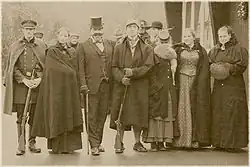 Das sepiafarbene Foto zeigt sieben Personen, die sich für ein Gruppenfoto in einer Reihe aufgestellt haben; die Frauen tragen lange Röcke oder Kleider und Mäntel, die Männer Anzüge, Mäntel oder militärische Uniformen.