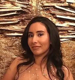 Latifa bint Muhammad Al Maktum mit lächelndem Gesichtsausdruck mit langen, dunklen Haaren, die ein durchsichtiges Oberteil trägt, das mit kleinen floralen Designs verziert ist. Die Wand im Hintergrund besteht aus einer kunstvollen, strukturierten Oberfläche mit horizontalen und diagonalen metallischen Elementen, die an abstrakte Kunst oder eine dekorative Wand erinnern.