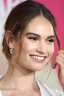 Lily James bei der Premiere des Films Baby Driver im Jahr 2017. Sie trägt ein elegantes weißes Outfit und hat ihr braunes Haar in einer lockeren Hochsteckfrisur gestylt. Ein besonderes Detail sind ihre Ohrringe: Am linken Ohr trägt sie zwei Creolen, eine davon mit einem kleinen Stern- und Perlenanhänger. Ihre Hand ist nahe an ihrem Gesicht, während sie eine Haarsträhne hält. Der Hintergrund ist in einem kräftigen Pink gehalten.