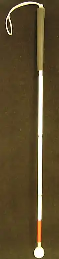 Ein weißer Langstock. Er hat am oberen Ende einen bräunlichen Griff und eine Handschlaufe, am unteren Ende eine orangefarbene Markierung und eine Kugel von der Größe eines Golfballes.