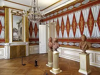 Farbige Eckansicht eines Innenraums mit drei Büsten, die im Raum auf Podesten stehen. Die roten Wände bestehen aus nebeneinander gemalten, weißen Vorhängen, die in der Mitte mit Kordeln gebunden sind. Zwischen den Vorhängen sind braune Ornamente. An der rechten Wand steht eine Flügeltür offen und an der linken Wand befindet sich ein vertikales Gemälde über einem Kamin mit goldenen Gittern davor. Ein Kronleuchter hängt von der Decke.