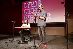 Man sieht zwei Männer auf einer Bühne, der eine sitzt an einem kleinen Holztisch, der andere steht. Beide haben ein Standmikorofon vor sich. Hinter ihnen ist auf einem rötlichen Hintergrund der Schriftzug „Lyrik geht weiter“ zu lesen.