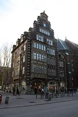 Seitliche Farbfotografie eines mehrgeschossigen, dunklen Gebäudes mit einem Geschäft im Erdgeschoss. Die linke Fassade hat über drei Stockwerke Fenstervorbauten und ein Dachgiebel schließt die Front oben ab. Rechts ist ein Gebäudeteil mit einem Dachkreuz zu sehen.