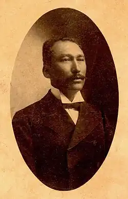 Mann im dunklem Anzug mit weißen Hemd und schwarzer Fliege ab Brustkorb von leicht seitlich vorn stehend leicht nach rechts schauend.