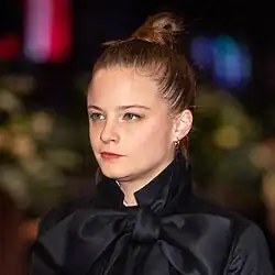 Jasna Fritzi Bauer auf der Berlinale 2020, trägt einen schwarzen Mantel mit großem Schleifenkragen, hat einen ernsten Gesichtsausdruck.Ein besonderes Detail ist die große, auffällige Schleife am Kragen, die aus einem glänzenden, satinartigen Material besteht und sehr sorgfältig gebunden ist. Die Frisur ist ein hoher Dutt, und als Accessoires trägt die Person kleine Kreolen-Ohrringe. Der Hintergrund ist unscharf, mit erkennbaren grünen Elementen – möglicherweise Pflanzen oder Bäume.