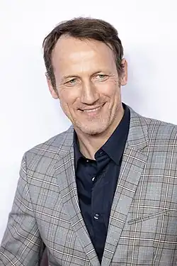 Wotan Wilke Möhring beim Berlinale-Empfang der nordrhein-westfälischen Landesvertretung in Berlin im Jahr 2020. Er trägt ein kariertes Sakko in Grau- und Blautönen, kombiniert mit einem dunkelblauen Hemd. Er steht vor einem neutralen Hintergrund.