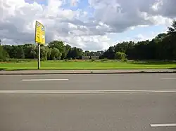 Im Vordergrund befindet sich eine mehrspurige Straße. Hinter dieser fällt die Höhe auf die Ebene der Wiese, die von Bäumen gesäumt ist.