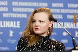 Maria Dragus bei der Pressekonferenz zum Film Tiger Girl auf der Berlinale 2017. Sie trägt ein schwarzes Oberteil mit weißen Sternen und große goldene Ohrringe, hat lange, gewellte blonde Haare und schaut nach rechts. Vor ihr ist ein Mikrofon platziert, was darauf hinweist, dass sie spricht oder gleich sprechen wird. Im Hintergrund befindet sich eine blaue Wand mit verschiedenen weißen Schriftzügen, darunter „BERLINALE“ und Begriffe wie „KULINARISCHES KINO“, „GENERATION“, „WETTBEWERB“, „NATIVE“, „EUROPÄISCHES KINO“, „PERSPEKTIVE DEUTSCHES KINO“, „SPECIAL“ und „PANORAMA“. Zudem sind Logos von Sponsoren wie L’Oréal und Audi zu erkennen.