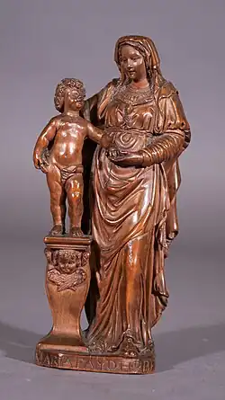 eine kleine rötlich-braune Buchsbaumfigur einer Madonna mit Kind. Das nackte Jesuskind steht auf einem Podest und reicht Maria bis zur Schulter. Am Fuß ist eine Signatur zu erkennen