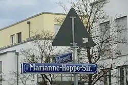 Blaues Straßenschild mit der Schrift "Marianne-Hoppe-Straße" am selben Ständer wie das schwerer lesbare, nach hinten wegstehende ebensolche Schild "Federseestraße" und darüber die Rückseite eines dreieckigen Verkehrsschildes. Dahinter unscharf moderne Wohngebäude und Bäume.
