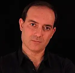 Massimo Maugeri (2013) trägt ein schwarzes Hemd und hat dunkles, kurzes Haar. Der Hintergrund des Bildes ist vollständig schwarz, wodurch er im Vordergrund besonders hervorgehoben wird. Es gibt keine weiteren sichtbaren Elemente oder Dekorationen im Bild.