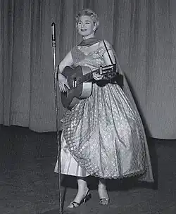 Eine Frau mit kurzen blonden Haaren in Tüllkleid und Stöckelschuhen, mit einem Schal um den Hals steht vor einem Mikrofon auf einer Bühne; sie spielt Gitarre und singt.