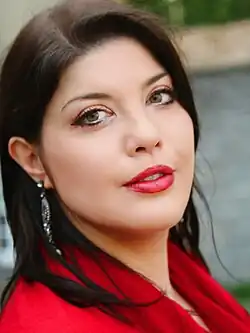 Frau mit langen, glatten, dunkelbraunen Haaren, die roten Lippenstift und roten Stoff um die Schultern trägt und mit leicht nach hinten geneigtem Kopf über ihre linke Schulter in die Kamera blickt.