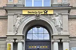 weibliche Sandsteinfiguren über dem Haupteingang, links mit Morsetaster, rechts mit aus einem Füllhorn fallenden Briefen.