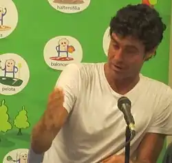 Miki Oca trägt ein weißes T-Shirt und sitzt vor einer grünen Wand mit Werbeaufschriften.