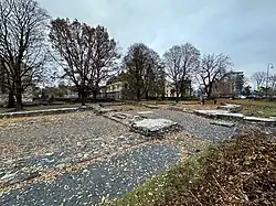 Foto einer Parkfläche mit Grundrissresten. Etwas entfernt befinden sich mehrstöckige Wohnblöcke.