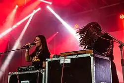 Beide Frauen sind auf der in rotes Licht getauchten Bühne zu sehen. Hélène de Thoury steht am Synthesizer mit wehenden Haaren. Die hinter einem Synthesizer stehende Amandine Stioui hält sich das Mikrophon vor den Mund und singt.