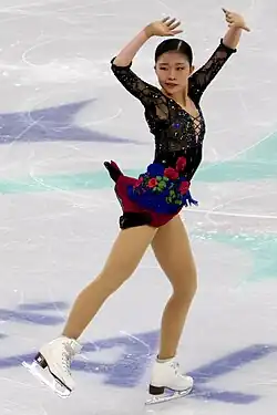 Mone Chiba in bewegter Pose mit dem Gewicht auf dem linken Bein, das rechte Bein nach hinten gestreckt, die Arme erhoben. Sie trägt ein schwarz-blaues Kleid mit roten Rosen.