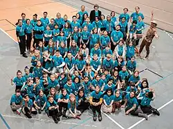 Etwa 100 Kinder des Musaik-Orchesters in blauen T-Shirts sind in einer Turnhalle sternförmig aufgestellt und schauen nach oben in die Kamera