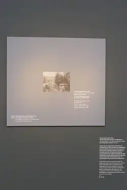 Braungraue Wand, vor der ein Rechteck in derselben Farbe in Größe des verliehenen impressionistischen Bildes hängt. Darauf ist klein mittig das verliehene Bild in Grautönen gedruckt, daneben Text auf Deutsch und Englisch