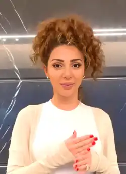 Myriam Fares mit lockigen Haaren, die zu einem hohen Dutt zusammengebunden sind. Sie trägt ein weißes Langarmshirt und darüber eine helle Strickjacke. Beide Hände der Person sind sichtbar, sie haben rote Fingernägel und scheinen eine Gestik mit beiden Händen zu machen. Der Hintergrund ist dunkel, möglicherweise eine Wand, mit einigen Lichtreflexionen und Streifen.