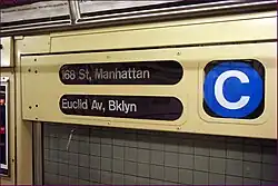 Detailansicht im Inneren eines U-Bahn-Wagens: Ein begie lackierter Metallkasten mit drei verglasten Öffnungen zeigt in gedrängter Form Informationen zum Zuglauf an. Links oben steht „168 St, Manhattan“, links unten steht „Euclid Av, Bklyn“, beides in weißer Schrift auf schwarzem Rollband. Rechts ist groß das runde blaue Liniensignet mit einem weißen Großbuchstaben C zu sehen.