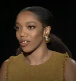 Naomi Ackie (2024) hat dunkle, lockige Haare, die nach hinten frisiert sind, und trägt große, goldene Kreolen-Ohrringe. Sie trägt ein ärmelloses Oberteil in Senfgelb. Das Oberteil hat eine strukturierte Oberfläche und ausgefranste Kanten an den Armlöchern und am Ausschnitt. Naomi Ackie schaut an der Kamera vorbei. Der Hintergrund ist vollständig dunkel.