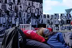 Eine Aktion der Betroffeneninitiative NichtGenesen am 15. März 2023 für Forschung vor dem Bundesministerium für Bildung und Forschung (BMBF). Im Vordergrund liegt eine Person in einem Bett. Die Person trägt eine Augenmaske. Im Hintergrund sieht man viele Schwarz-Weiß-Fotos von weiteren Personen, die an einem Zaun befestigt sind. Ein Kreuz symbolisiert anstelle eines Fotos eine verstorbene Person.