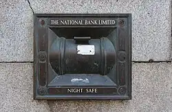 Ein Nachttresor ist in eine graue Steinwand eingelassen und ragt aus der Wand heraus. Er ist rechteckig und hat eine abgerundete, zylindrische Form. Die Oberfläche des Tresors ist dunkelgrau, wobei einige Gebrauchsspuren oder Abnutzungserscheinungen sichtbar sind. Auf dem Tresor sind die Worte „THE NATIONAL BANK LIMITED“ und „NIGHT SAFE“ lesbar.