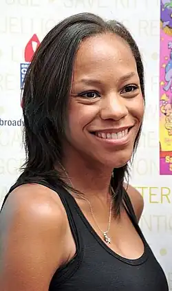 Nikki M. James nimmt 2011 am 13. jährlichen Broadway Barks Benefit in der Shubert Alley in New York City teil. Sie trägt ein ärmelloses schwarzen Oberteil und eine Halskette mit einem Anhänger. Der Hintergrund enthält Texte und Logos, darunter der Schriftzug „SAVE KITTY“ und „BROADWAY CARES“. Auf der rechten Seite ist ein farbenfrohes Poster zu sehen.